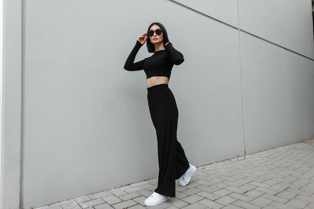 Fashionable beautiful girl in black stylish sportswear clothes\
with white sneakers walks near a gray wall and puts on cool trendy\
sunglasses in the city