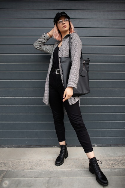 Modello alla moda bella ragazza in abiti casual alla moda con una camicia, jeans, una maglietta nera, scarpe con una borsa e un berretto si trova vicino a un cancello di metallo