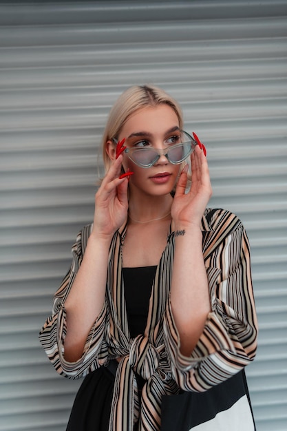 Fashionable beautiful girl in stylish clothes wears cool blue sunglasses near the metal wall on the street