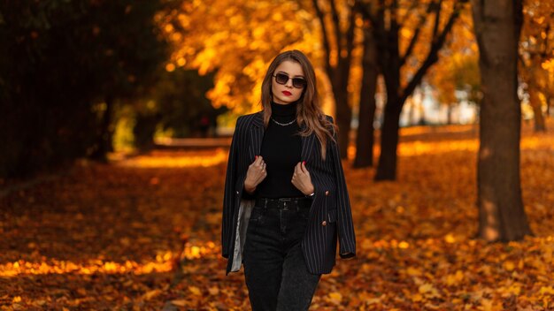 Bella ragazza alla moda con occhiali da sole vintage in abiti neri alla moda con un blazer alla moda, un maglione e jeans a piedi in un parco autunnale con fogliame autunnale arancione