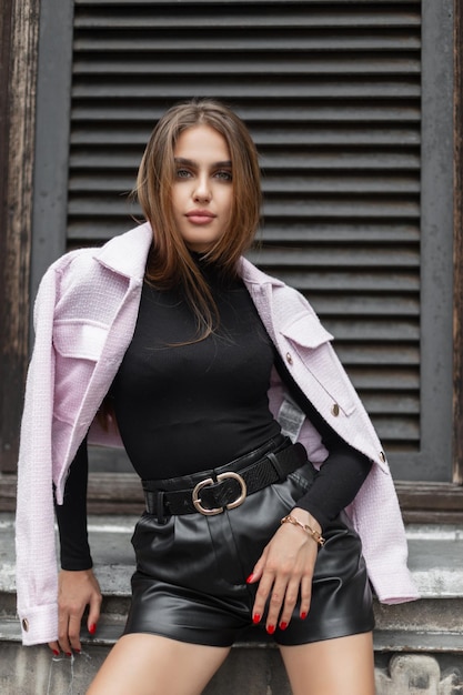 Fashionable beautiful glamor girl model in stylish elegant clothes with a black long sleeve shirt and leather shorts with a pink jacket on her shoulders near a vintage building