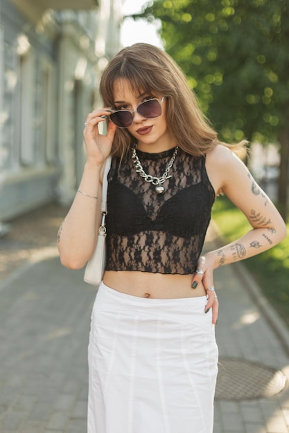 Fashionable beautiful hipster girl with a tattoo on arms in trendy fashion summer clothes with lace top white skirt with a handbag walks in the city and adjusts vintage cool sunglasses