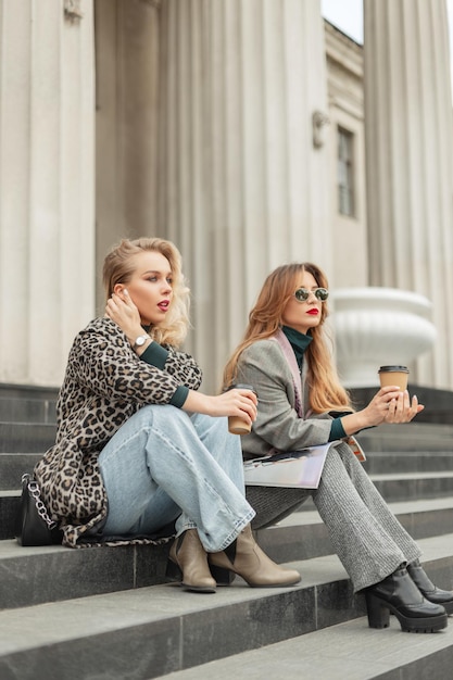 Fashionable beautiful model girl friends in stylish vintage clothing sitting outside a vintage building with columns and drinking coffee in the city