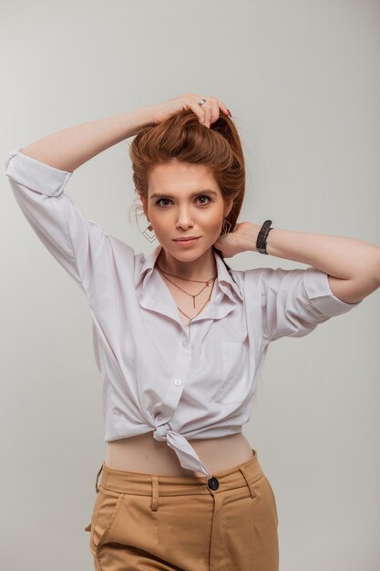 Fashionable beautiful redhead woman model with a clean face in a stylish white shirt in the studio on a gray background