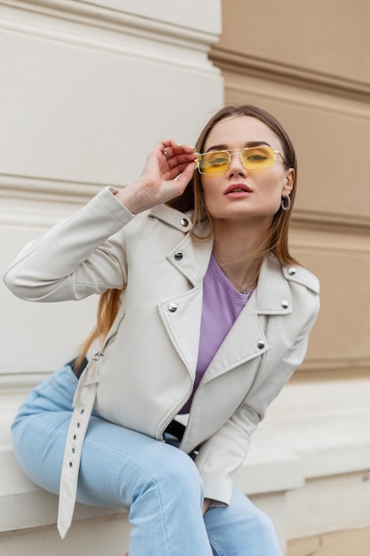 Fashionable beautiful stylish glamorous woman with vintage yellow sunglasses in a fashionable white leather jacket and blue jeans sits near a beige wall on the street