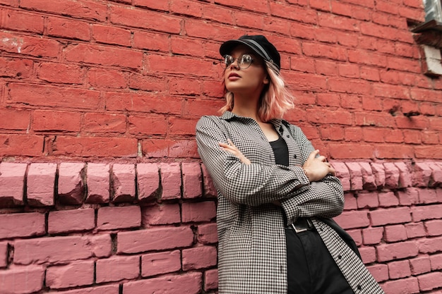 Bella ragazza alla moda hipster alla moda con gli occhiali e un berretto in abiti casual con una borsa si trova vicino a un vecchio muro di mattoni vintage