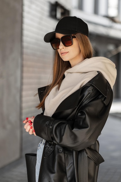 Fashionable beautiful stylish hipster girl with stylish vintage sunglasses and a black cap mockup wearing trendy urban clothing with a hoodie leather coat and bag walking on the street