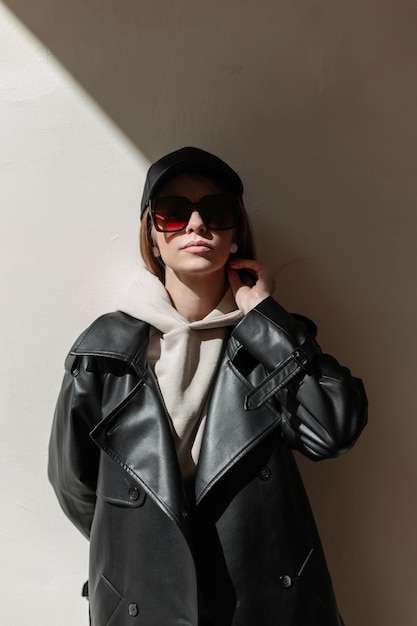 Fashionable beautiful stylish hipster woman with cool trendy sunglasses and a black cap in a fashionable leather coat with a hoodie stands in the sunlights near the wall on the street