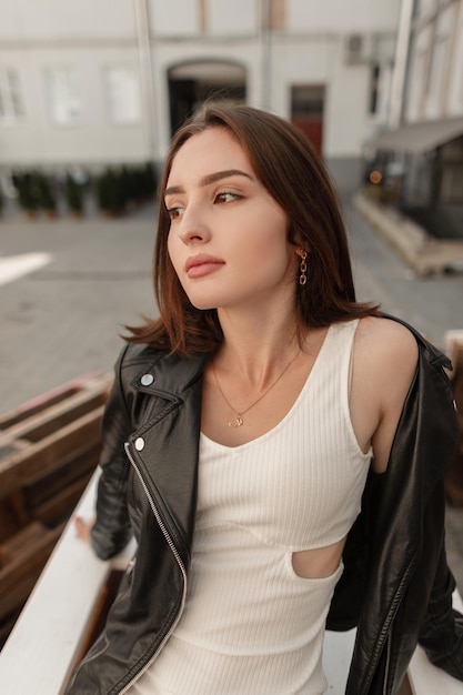 Fashionable beautiful stylish woman model hipster with short hair in a fashion black leather jacket with a white dress sits and relaxes in the city on wooden pallets