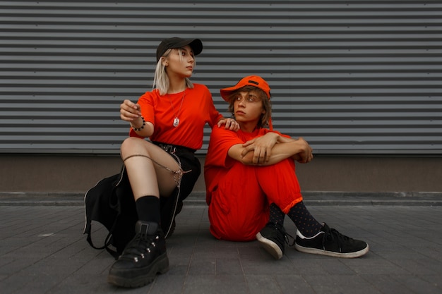 Фото Модная красивая стильная молодая пара в кепках в модной оранжевой одежде с модной обувью сидит на улице у металлической серой стены