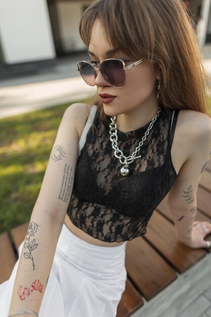 Fashionable beautiful urban hipster girl with vintage sunglasses with a tattoo on arms in a stylish lace top and a bra and a white skirt is sitting on a bench on the street