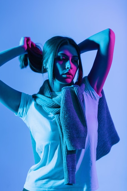 Fashionable beautiful woman in stylish casual clothes with white t-shirt and gray sweater on blue background with pink light