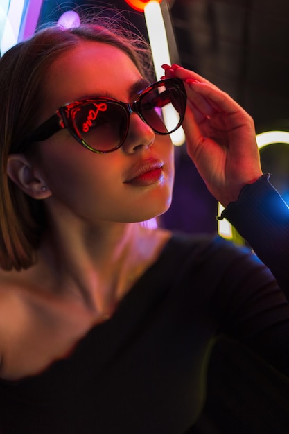 Fashionable beautiful woman in stylish clothes wears vintage sunglasses on a dark background with neon light Beauty style and fashion