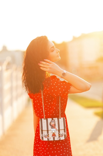 Foto la bella donna alla moda con capelli ricci in un vestito rosso con una borsa alla moda cammina sulla via al tramonto