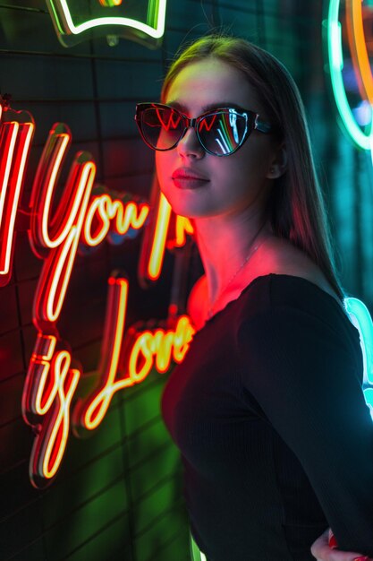 Fashionable beautiful woman with a fashion black dress and stylish sunglasses on a dark background with colored neon lights