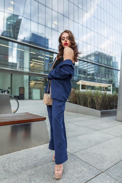 Fashionable beautiful young business woman with a hairstyle with red lips in a fashion blue suit with stylish shoes with a handbag walks in the city near office buildings pretty chic girl