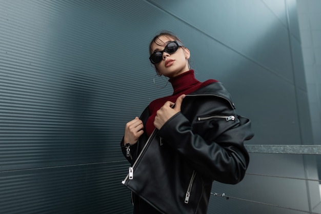 Fashionable beautiful young model woman in vogue sunglasses in stylish black leather jacket with knitted jersey posing near modern metal wall in the city