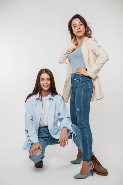 Модные красивые молодые женщины-подружки в стильной модной джинсовой одежде с блейзером, рубашкой, джинсами и каблуками позируют на белом фоне в студии