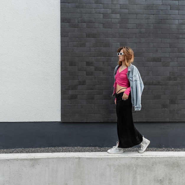 Модная красивая модель молодой женщины с крутыми белыми солнцезащитными очками в модной одежде с розовой верхней джинсовой курткой и юбкой с кроссовками, идущими возле кирпичной черной стены на улице