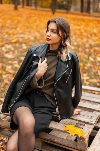 Fashionable beautiful young woman model with a fashion leather jacket and a vintage sweater with shorts sits on a wooden pallet in an autumn golden park