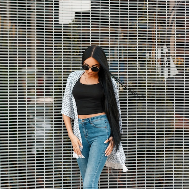 Fashionable beautiful young woman in stylish round sunglasses in a white cape stands near the wall