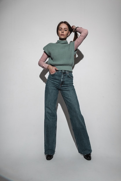 Photo fashionable beautiful young woman in a stylish vintage outerwear with knitted green vest pink shirt and flared jeans in the studio glamorous girl in retro look womens flared and wide leg 70s style