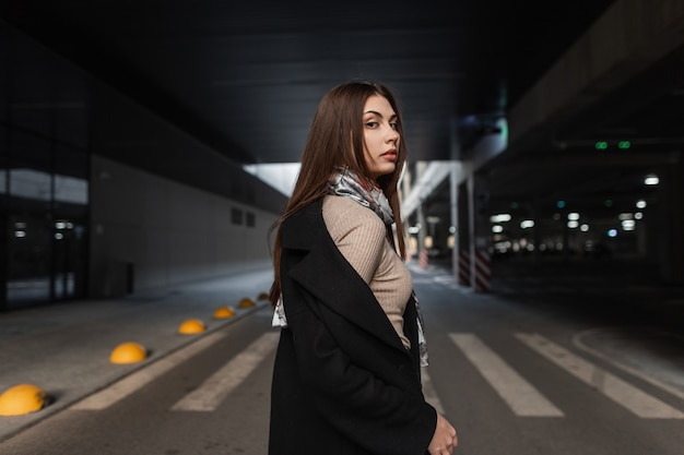 Fashionable beautiful young woman with brown hair in black fashionable coat in shirt stands on the road in the street. Pretty modern luxurious girl fashion model walks outdoors. Trendy lady.