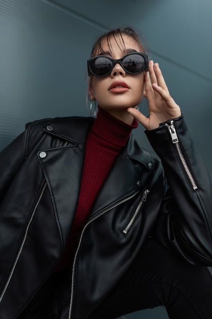 Fashionable beautiful young woman with cool sunglasses in stylish black leather jacket with burgundy sweater posing in the city