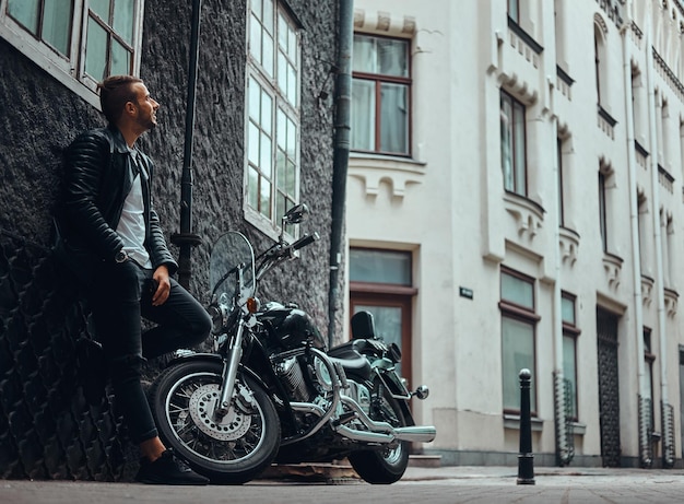 Fashionable biker dressed in a black leather jacket and jeans leaning on a wall near his retro motorcycle on an old Europe street.