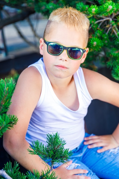 Fashionable blond boy in dark glasses. around a pine branch