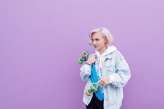 Foto ragazza bionda alla moda in una giacca di jeans si erge con uno skateboard sullo sfondo di un muro viola e si affaccia sul lato. copyspace