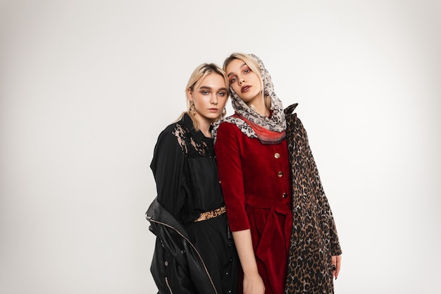 Fashionable blond woman in stylish oversized youth black jacket in elegant gloves and girl fashion model with scarf on head in luxurious leopard coat poses indoors