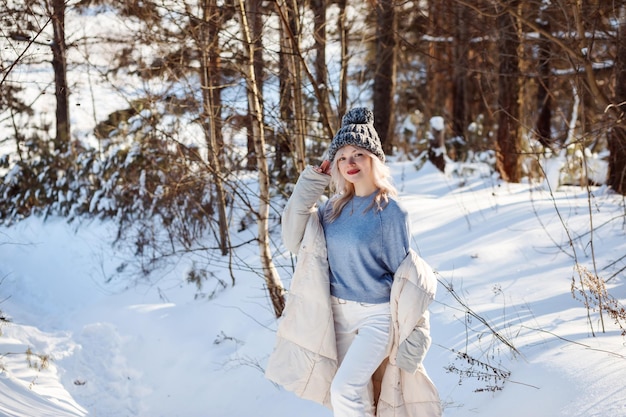 冬の森のクリスマスに屋外で暖かいキャップの青いセーターとベージュのコートでファッショナブルなブロンドの女の子...