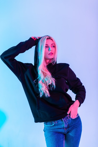 Fashionable blonde girl with a black sweatshirt wears a hoodie in blue jeans in the studio on a colored neon pink background