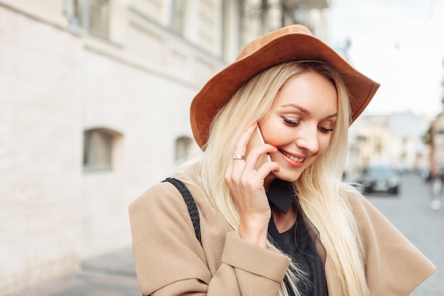 Fashionable blonde woman in felt hat talking on the phone. Portrait of cute girl in the city