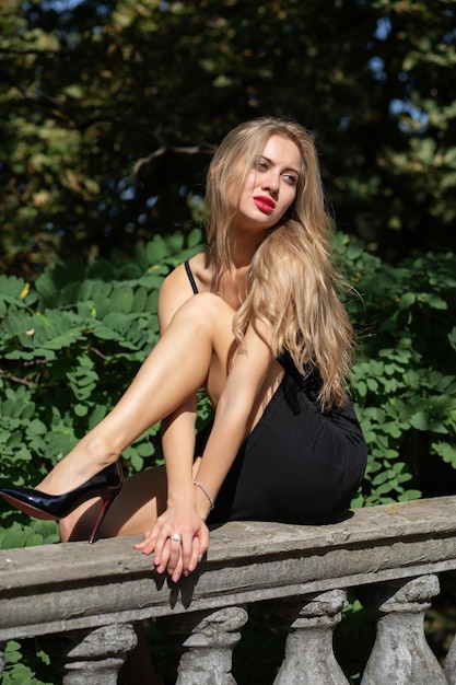 Модная блондинка с пышными волосами в классическом черном платье и туфлях сидит на балясинах