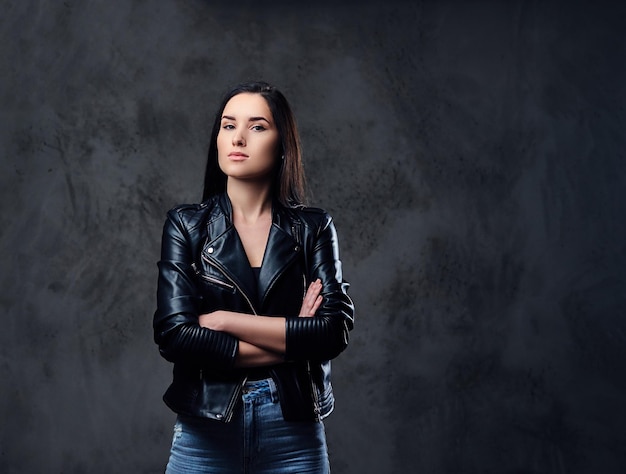 A fashionable brunette female dressed in a black leather jacket and a jeans over dark grey background.