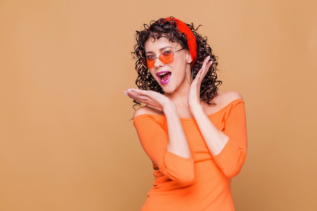 Fashionable brunette girl in a bright orange sweater, glasses and a bandana posing on orange background