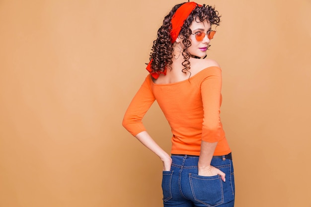 Модная брюнетка в ярко-оранжевом свитере, очках и бандане позирует на оранжевом