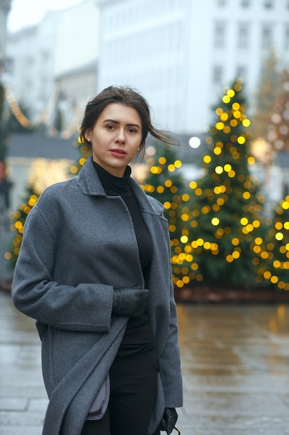 Fashionable brunette woman wears trendy coat walking in a rainy day at the city decorated to Christmas