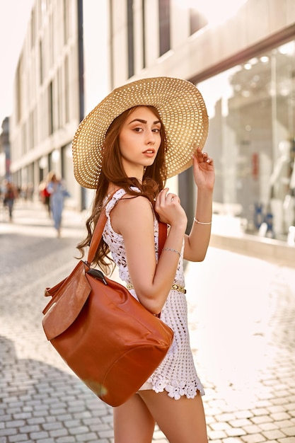 Fashionable brunette woman in a white dress straw hat walking in the street posing near shop windows