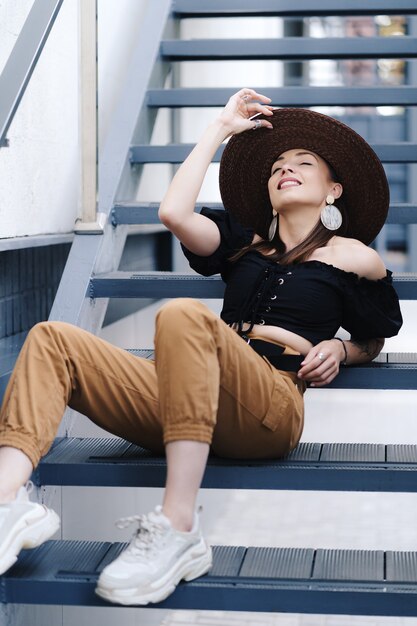 Fashionable brunette woman with long hair, wearing stylish large wicker hat, posing on the stairs.