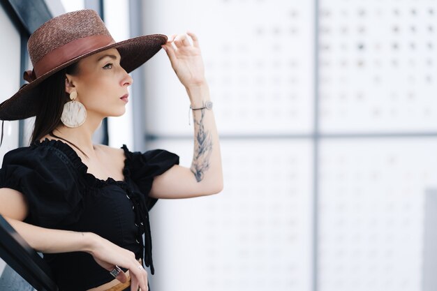 Fashionable brunette woman with long hair, wearing stylish large wicker hat, posing on the stairs.