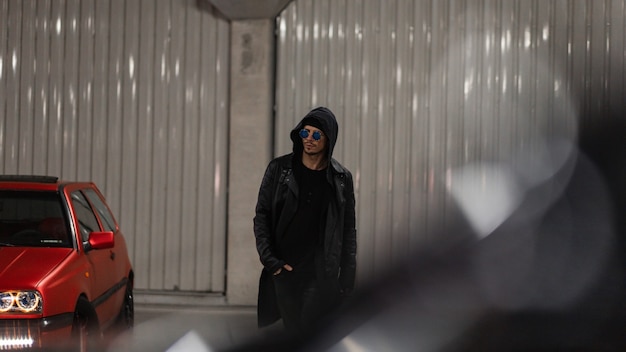 Photo fashionable brutal man model with hooded with sunglasses in a stylish black leather jacket with a hoodie walks in the parking lot at night