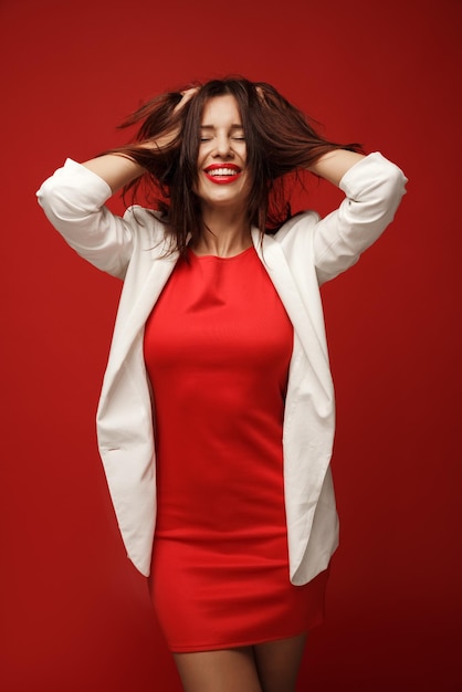 Fashionable cheerful girl in a red dress and a white jacket shatters her hair isolated on a red background