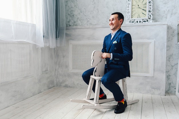 Fashionable cheerful man in a blue suit is sitting on a toy horse. 