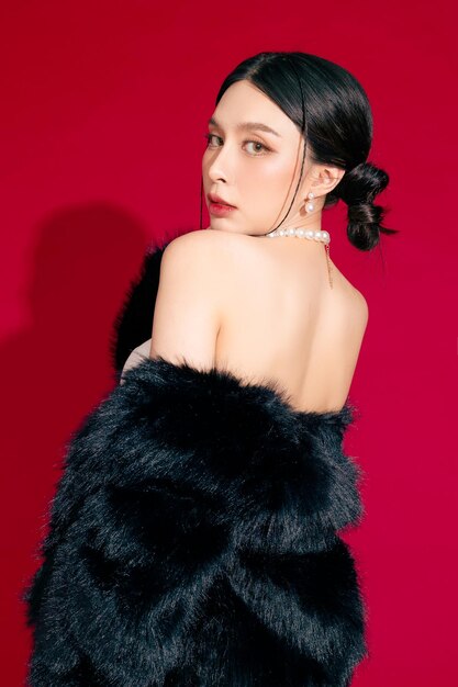 Fashionable confident asian woman wearing chic fur jacket and black boots in perfect slim shape on isolated red background in studio cosmetics femininity