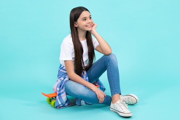 Photo fashionable confident teenage girl wearing trendy tshirt and jeans sit on skateboard posing on blue background fashion teenager happy teenager positive and smiling emotions of teen girl