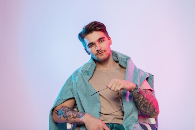 Fashionable cool handsome man model artist with hairstyle and stylish vintage eyewear in casual trendy outfit with tattoos on arms sits in studio with colorful pink blue lights