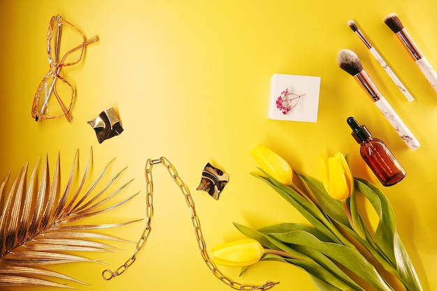 Fashionable cosmetics accessories makeup brushes and accessories on yellow background Palm leaves tulips glasses serum dropper decorations Copy space High quality photo